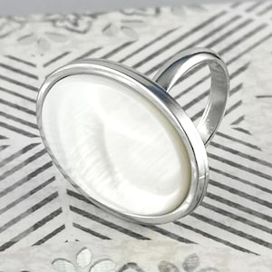 Könnte beinhalten: Ein silberfarbener Ring mit einer großen, kreisförmigen Einlage aus irisierendem weißem Perlmutt, die in eine glatte Metallfassung eingefasst ist. Die Ringschiene ist dünn und poliert und bietet eine saubere, reflektierende Oberfläche, die das leuchtende, perlmuttartige Zentrum der runden Fassung ergänzt.