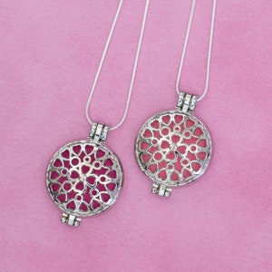 May include: Two silver-colored pendant necklaces with a round, openwork design featuring heart shapes. The pendants have a pink interior and hang from a thin silver chain. The background is a solid pink color.
