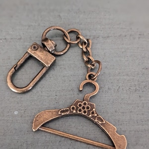May include: A bronze-colored metal keychain with a small, detailed hanger charm. The hanger has a floral design and a small bar across the top.