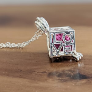 May include: A silver necklace with a charm that is a cube with the word "LOVE" spelled out on it. The cube is open and has a pink interior.