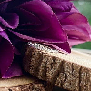 May include: A delicate silver band ring with a row of small, clear gemstones set in a bezel setting. The ring is resting on a piece of wood with a purple flower in the background.