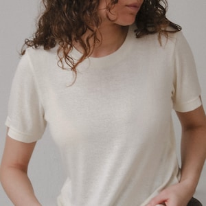 May include: A woman wearing a white, short-sleeved, crew neck t-shirt. She is pulling the bottom of the shirt up to show the hem.
