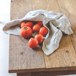 Könnte beinhalten: Eine Gruppe roter Tomaten fällt aus einer hellgrauen Leinentasche auf einen rustikalen Holztisch.