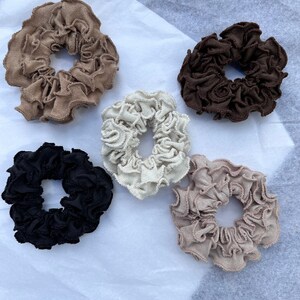 May include: Five fabric scrunchies in different colours: brown, black, beige, light brown, and white. The scrunchies are all ruffled and have a gathered look.