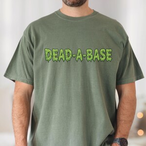 May include: A moss green t-shirt with the text "DEAD-A-BASE" in a dripping green font. The shirt is a short-sleeved, crew neck style. The model is wearing blue jeans and a black watch.