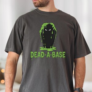 May include: Dark gray t-shirt featuring a black coffin design with green slime and the text "DEAD-A-BASE" in a dripping font. The shirt is a casual style, suitable for everyday wear.