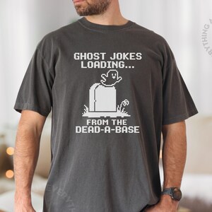 May include: Dark gray t-shirt with a white graphic that reads "GHOST JOKES LOADING... FROM THE DEAD-A-BASE". The design features a pixelated ghost and tombstone. The shirt is a comfortable, casual style.