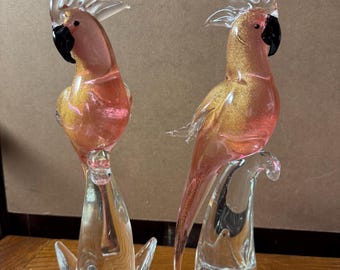 Peut inclure: Deux figurines de perroquets en verre. Les oiseaux ont des corps roses et dorés, des becs noirs et des crêtes transparentes. Ils sont perchés sur des socles en verre transparent. Les figurines sont exposées sur une surface en bois.