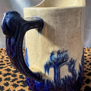 May include: Cream-colored ceramic pitcher with a blue handle and base. The pitcher features a blue landscape design around the base and a crackled glaze finish. The handle is a dark blue, with a unique, sculpted design.