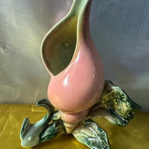 May include: A ceramic vase shaped like a conch shell with a glossy pink exterior and a pale green interior. The base features sculpted green and brown leaf-like accents that extend outward, providing a stable foundation for the upright shell form.