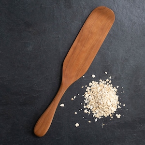 May include: A wooden spatula with a long handle and a wide, flat blade. The spatula is a warm brown color and is next to a pile of uncooked oatmeal. The background is a dark gray, textured surface.
