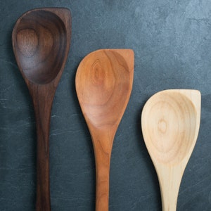 May include: Three wooden spoons of varying shades of brown, from dark to light. The spoons have a unique, asymmetrical design with a rounded bowl and a flat, angled edge. The spoons are arranged vertically against a dark gray background.