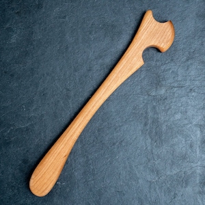 Puede incluir: Utensilio de cocina de madera clara. Tiene un mango largo y curvado y una cabeza con una forma distintiva, que incluye una muesca y un borde curvado. Este utensilio probablemente se utiliza para cocinar o servir alimentos.