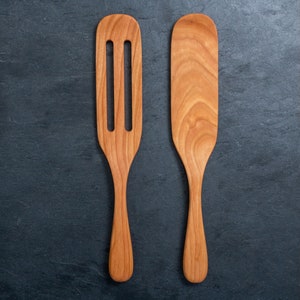 May include: Two wooden kitchen utensils: a slotted spatula and a solid spatula. Both are made of light brown wood with visible grain patterns. The slotted spatula has three rectangular cutouts. The utensils are designed for cooking and food preparation.