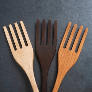 Puede incluir: Tres tenedores de ensalada de madera en tonos marrones y beige. Los tenedores tienen cuatro púas y están colocados sobre un fondo gris oscuro. La veta de la madera es visible en cada tenedor, resaltando el material natural.