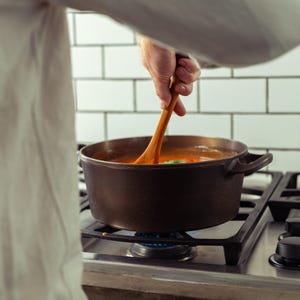 May include: A dark brown cast iron pot filled with a red liquid, possibly soup or stew, is on a gas stove. A wooden spoon stirs the contents. The stove is black, and the background features white tiles.