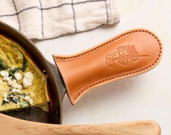 May include: A tan leather handle cover for a cast iron skillet, featuring a debossed horse-drawn carriage logo and stitched edges. The cover is fitted onto the handle of a dark metal pan containing a cooked egg dish with spinach and white cheese.