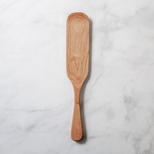 May include: A light-colored wooden spatula with a long, rectangular head and a slender handle. The spatula is made of a light-colored wood, and the grain is visible. The spatula is designed for cooking and food preparation.
