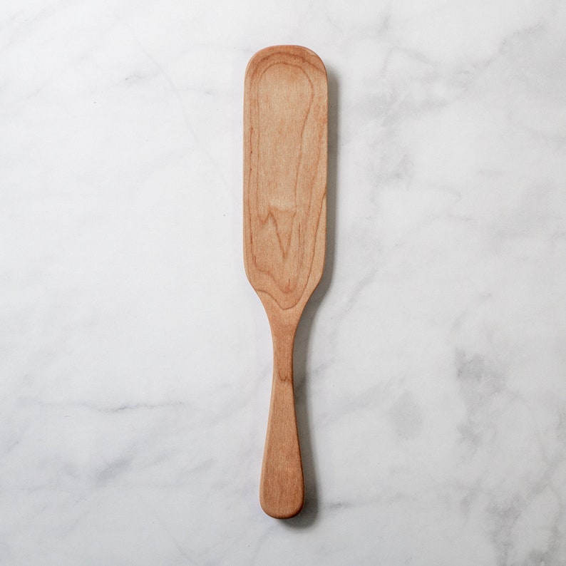 May include: A light-colored wooden spatula with a long, rectangular head and a slender handle. The spatula is made of a light-colored wood, and the grain is visible. The spatula is designed for cooking and food preparation.
