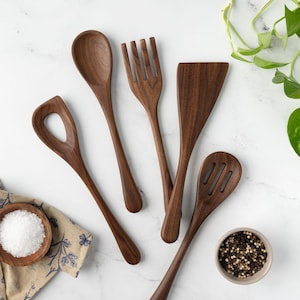 May include: A set of five dark brown wooden kitchen utensils including a solid spoon, a four-tined fork, a flat spatula, a slotted spoon, and a spoon with a large central hole, arranged on a white surface with a small bowl of salt and pepper.