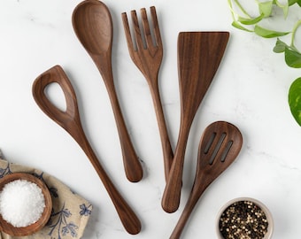 Puede incluir: Un juego de cinco utensilios de cocina de madera de color marrón oscuro que incluye una cuchara sólida, un tenedor de cuatro dientes, una espátula plana, una cuchara ranurada y una cuchara con un gran orificio central, sobre una superficie blanca.