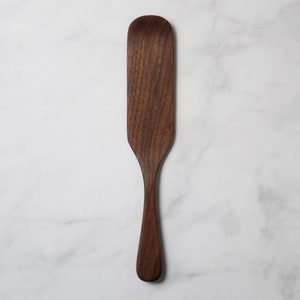 May include: A handcrafted wooden spatula with a long handle and a wide, rounded head. The spatula is made of dark brown wood and has a smooth, polished finish. The kitchen utensil is designed for cooking and serving.