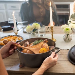May include: A cast iron pot filled with roasted chicken, potatoes, and lemon slices. A wooden spoon rests inside. The pot is on a wooden table, with lit candles and flowers in the background. A person is holding the pot.