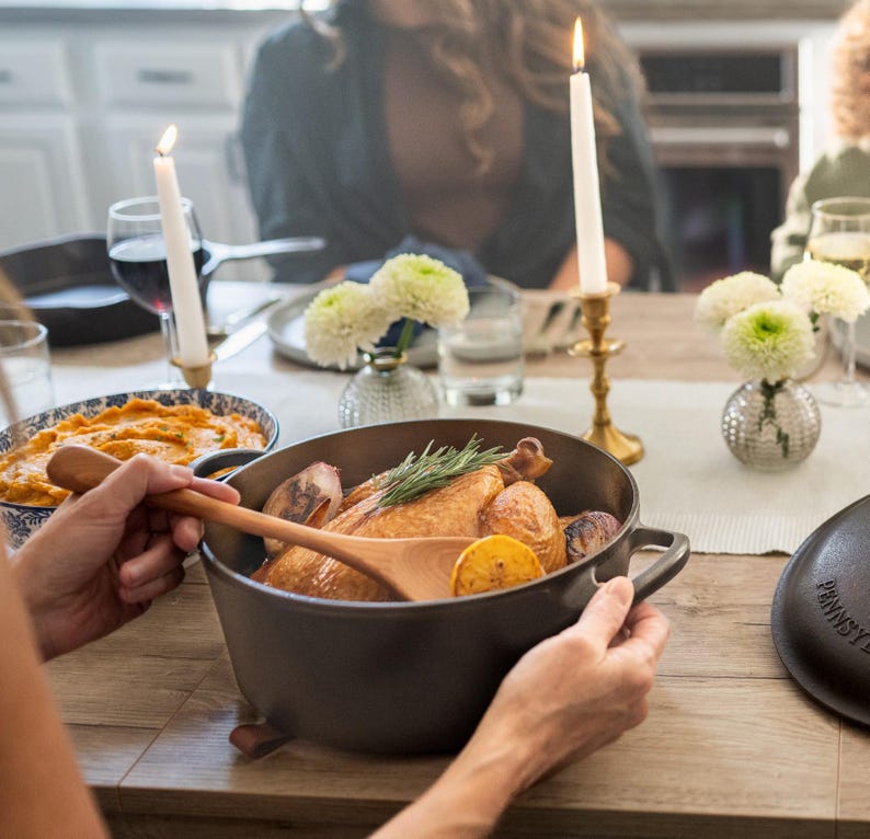 May include: A cast iron pot filled with roasted chicken, potatoes, and lemon slices. A wooden spoon rests inside. The pot is on a wooden table, with lit candles and flowers in the background. A person is holding the pot.