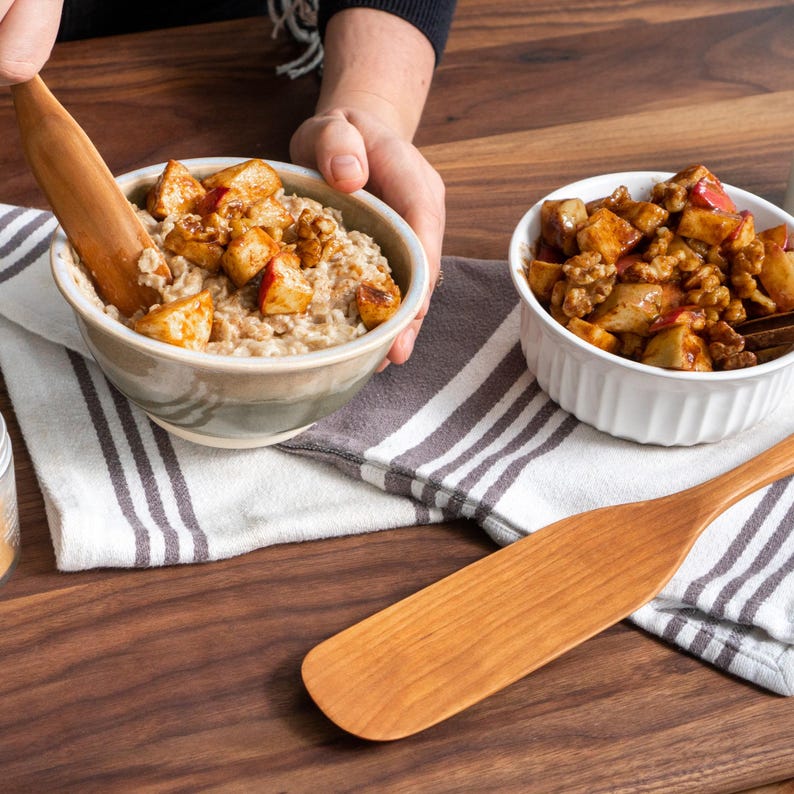 May include: Two bowls of oatmeal topped with diced apples and walnuts. One bowl is being stirred with a wooden spoon. The bowls are on a striped towel and a wooden surface.