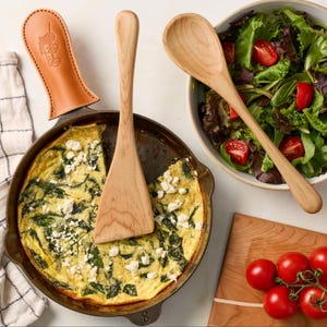 Op de afbeelding: Een gietijzeren koekenpan met een spinazie- en feta-frittata met een beige lederen handgreep, een houten spatel, een houten opscheplepel in een kom met gemengde sla en kerstomaatjes, en een houten snijplank met kerstomaatjes.