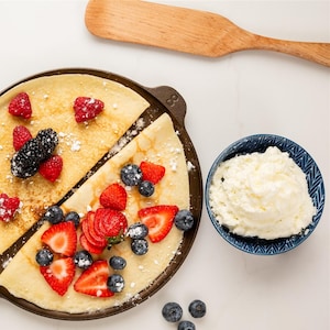 May include: A crepe pan with two crepes, one half topped with raspberries and blackberries, the other with sliced strawberries and blueberries. A small blue bowl of whipped cream and a wooden spatula are also in the image.