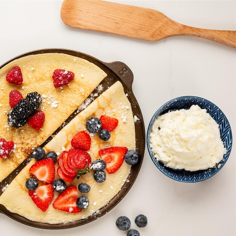 May include: A crepe pan with two crepes, one half topped with raspberries and blackberries, the other with sliced strawberries and blueberries. A small blue bowl of whipped cream and a wooden spatula are also in the image.