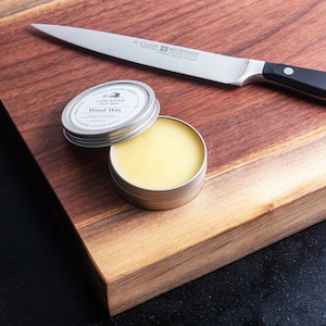 May include: A small tin of Lancaster Cast Iron Wood Wax, open and partially filled with a yellow wax, on a wooden chopping board with a silver knife.