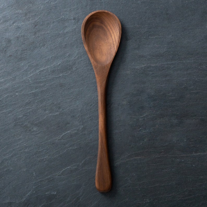 May include: A wooden spoon with a long handle and a rounded bowl. The spoon is made of a dark brown wood and is set against a dark gray background. The spoon is approximately 12 inches long.