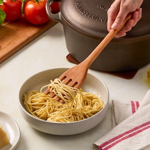 Puede incluir: Un tenedor para pasta de madera de color marrón claro con tres dientes que levanta una porción de espaguetis cocidos de un cuenco de cerámica beige moteado. El utensilio tiene un mango largo y una cabeza ranurada para servir pasta.