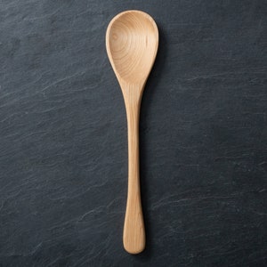 May include: A light-colored wooden spoon with a rounded bowl and long handle. The spoon is made of a light-colored wood and is set against a dark, textured background. The spoon is designed for cooking and serving.