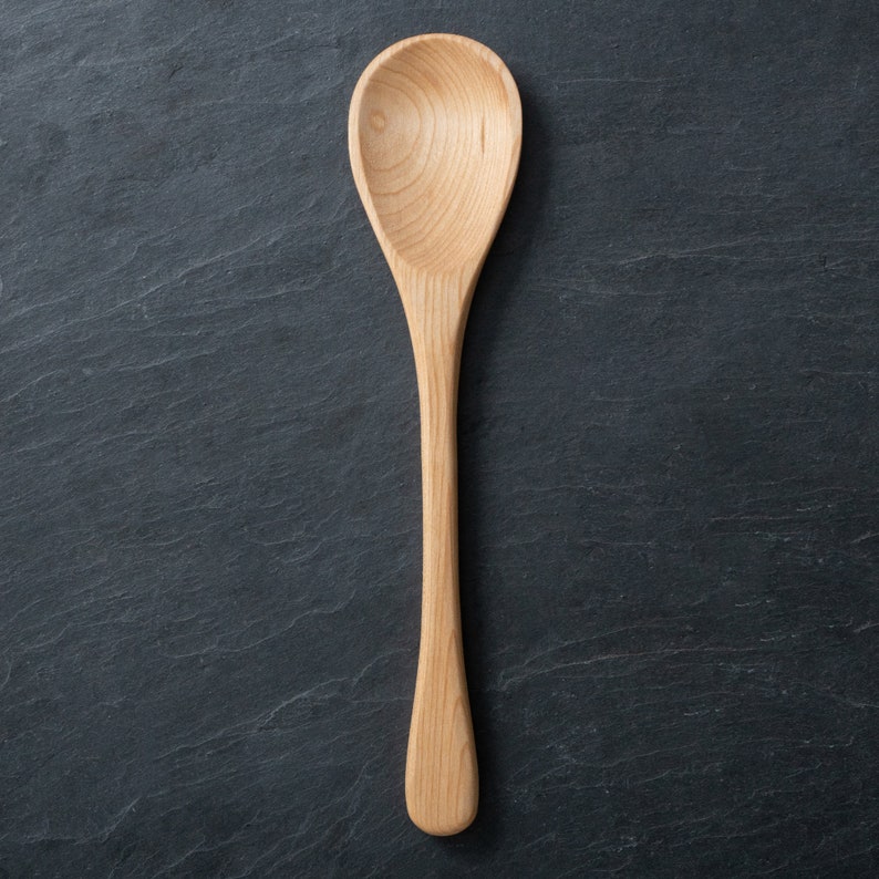 May include: A light-colored wooden spoon with a rounded bowl and long handle. The spoon is made of a light-colored wood and is set against a dark, textured background. The spoon is designed for cooking and serving.