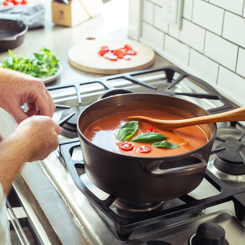 May include: A cast iron pot filled with tomato soup, garnished with basil leaves and sliced tomatoes, sits on a gas stove. A wooden spoon rests in the soup. The scene suggests home cooking.