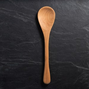 May include: A wooden spoon with a long handle and a rounded bowl. The spoon is made of light-colored wood and is set against a dark, textured background. The spoon is a kitchen utensil.