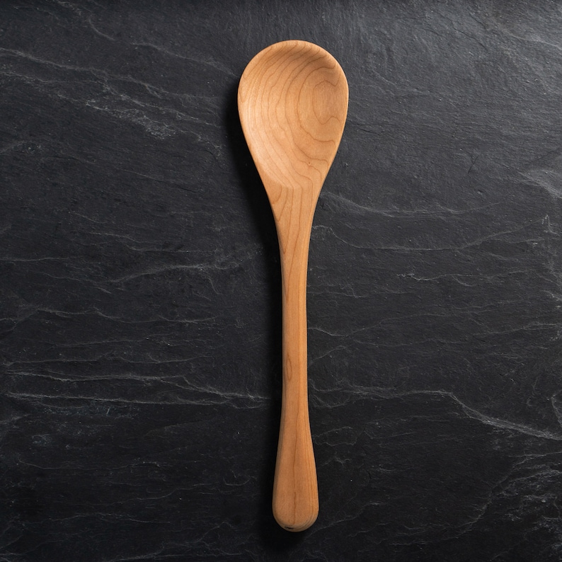May include: A wooden spoon with a long handle and a rounded bowl. The spoon is made of light-colored wood and is set against a dark, textured background. The spoon is a kitchen utensil.