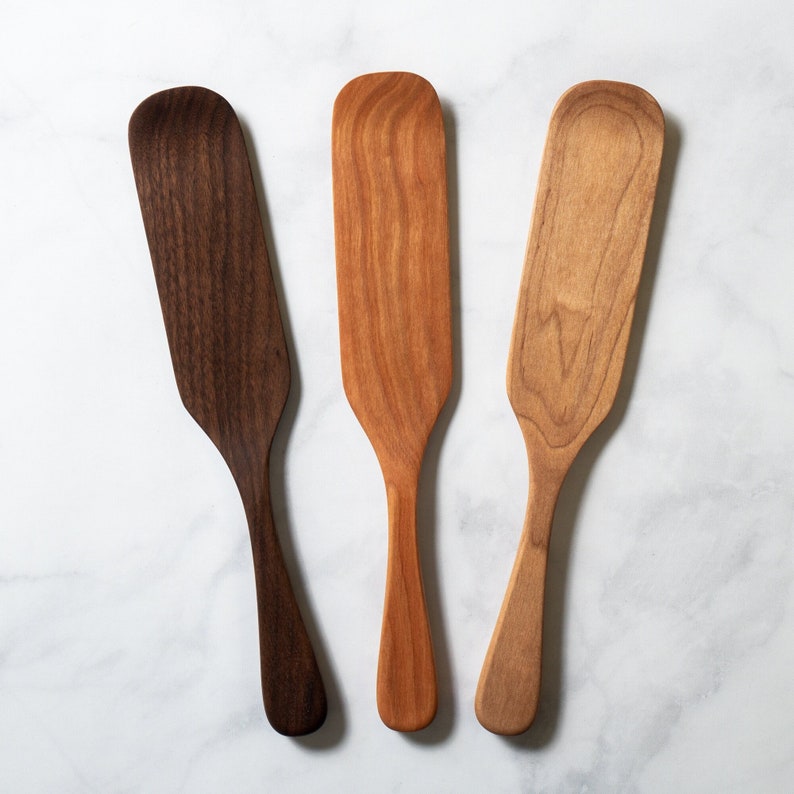 May include: Three wooden butter spreaders in varying shades of brown. The spreaders have a paddle-like shape with a rounded top and a long, slender handle. The wood grain is visible, adding to the natural aesthetic. The spreaders are arranged on a white marble surface.