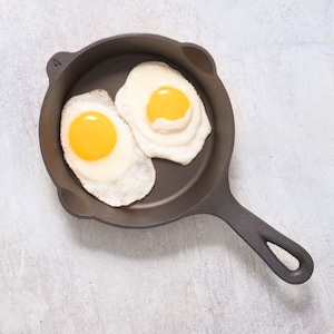 May include: Two fried eggs in a black cast iron skillet. The eggs are cooked sunny side up with the yolks still runny.