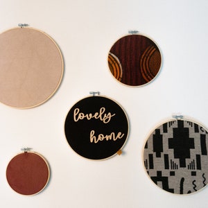 Könnte beinhalten: Fünf runde Stickrahmen mit verschiedenen Stoffdesigns. Die Rahmen hängen an einer weißen Wand. Ein Rahmen hat die Worte "lovely home" aufgestickt.