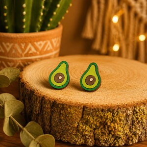 Könnte beinhalten: Ein Paar Ohrstecker in Form von Avocadohälften mit hellgrünem Fruchtfleisch, einem dunkelbraunen Kern und einer dunkelgrünen Schale. Die Ohrstecker sind aus Holz gefertigt und haben eine flache, bemalte Oberfläche mit sichtbarer Maserung.