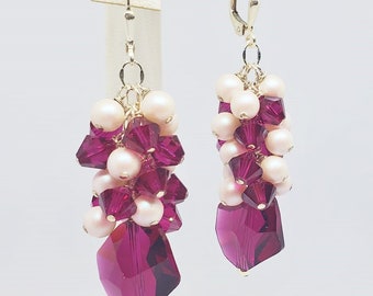 Peut inclure: Une paire de boucles d'oreilles pendantes avec un groupe de cristaux roses et fuchsia et de perles. Les boucles d'oreilles sont en métal argenté et ont une fermeture à crochet. "Glittering Ice Designs Handcrafted Artisan Jewelry" est écrit en bas de l'image.