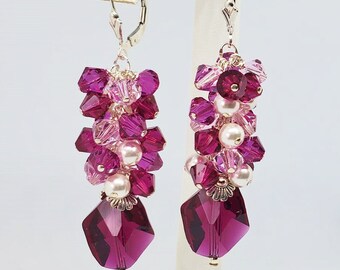 Peut inclure: Boucles d'oreilles pendantes composées de grappes de cristaux roses et magenta et de fausses perles. Les boucles d'oreilles ont un grand cristal magenta facetté en bas. Elles sont suspendues à des fermoirs argentés. Le texte "Glittering Ice Designs Handcrafted Artisan Jewelry" est en bas.