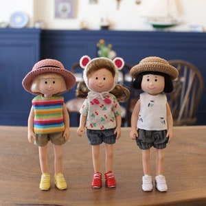 May include: Three handmade dolls dressed in colorful outfits. The doll on the left wears a pink hat, striped shirt, and shorts. The middle doll has a bear hat and cherry-print shirt. The right doll wears a straw hat and a white top with a blue design.