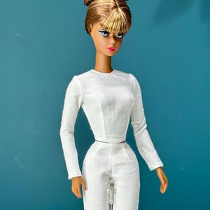 May include: A fashion doll with brown and blonde hair in a bun and bangs, wearing a white long-sleeved top and matching trousers. The doll has a slender waist and is posed against a teal background.