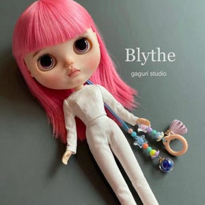 May include: A Blythe doll with pink hair and large eyes, wearing a white jumpsuit. The doll is accessorized with a beaded charm bracelet featuring stars and a ring. The text "Blythe" and "gaguri studio" are visible.