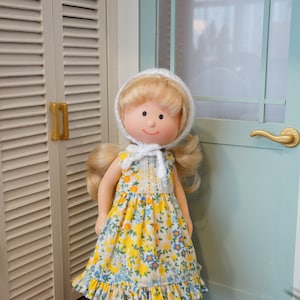 May include: A vintage doll with blonde hair and a white bonnet stands in front of a door. The doll wears a yellow floral print dress with a white lace collar and black shoes. The background includes a wooden floor and a light blue door.